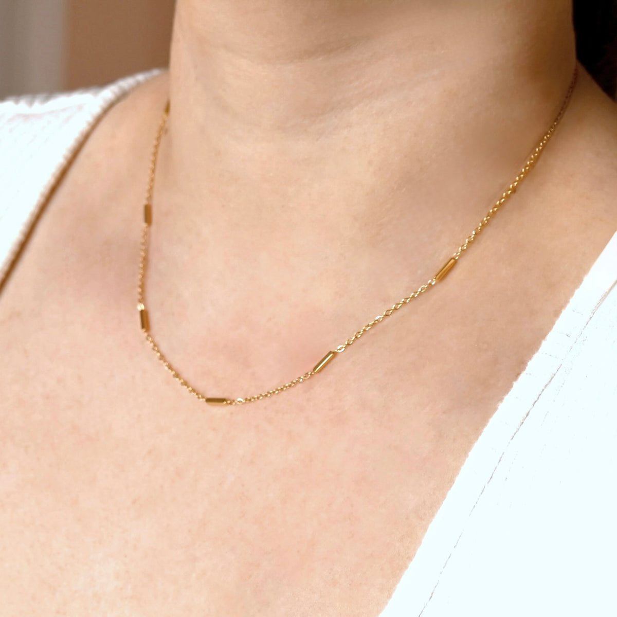 A delicate gold chain necklace with small polished bar links, worn on a model's neck over a white top.
