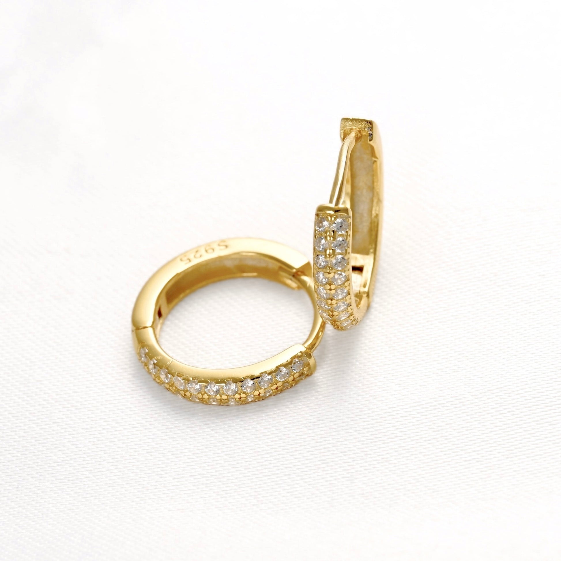 A pair of gold huggie hoop earrings with pavé-set clear stones, shown on a clean white textured background.