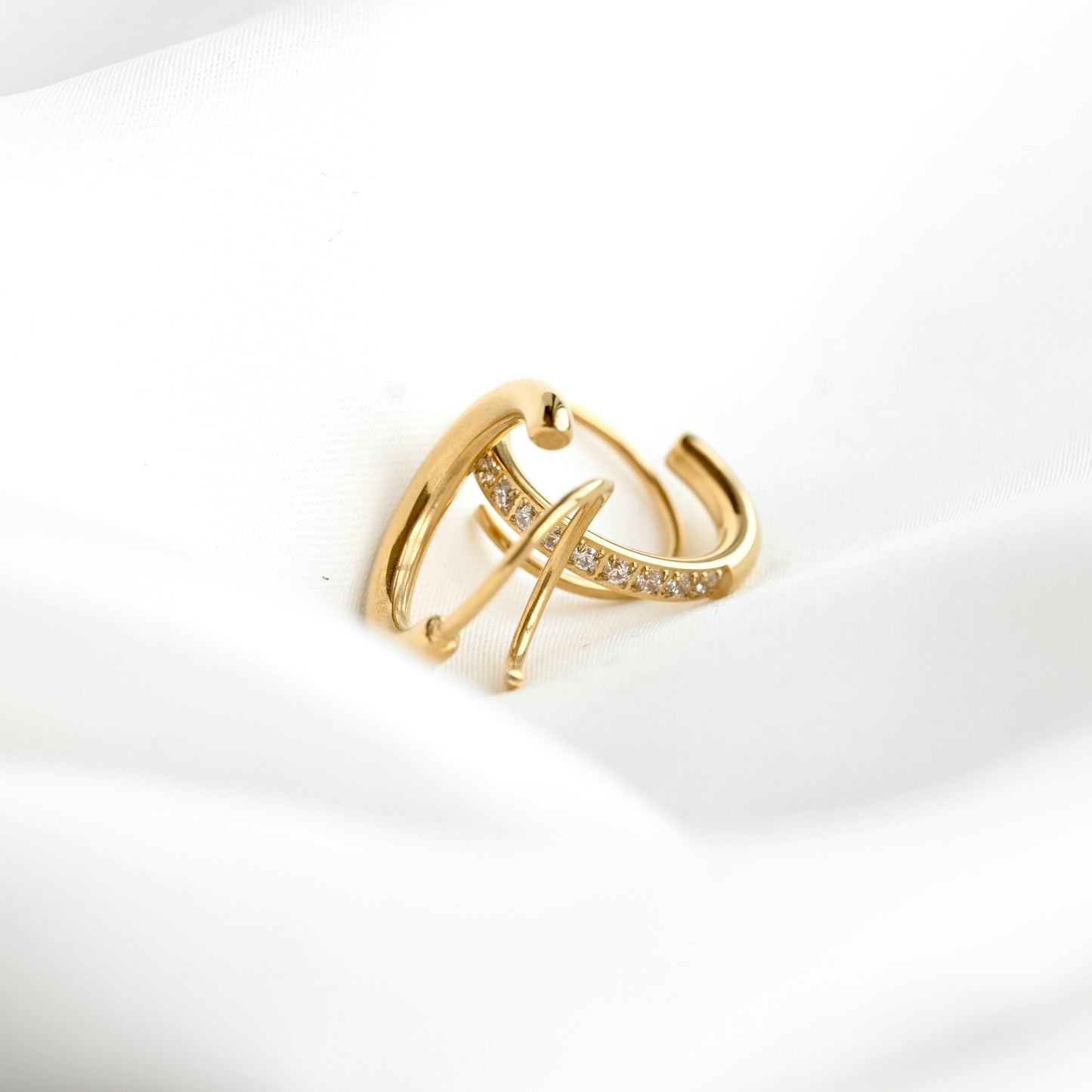 A gold huggie hoop ring with a curved bar studded with small clear crystals, resting on a soft white fabric background.
