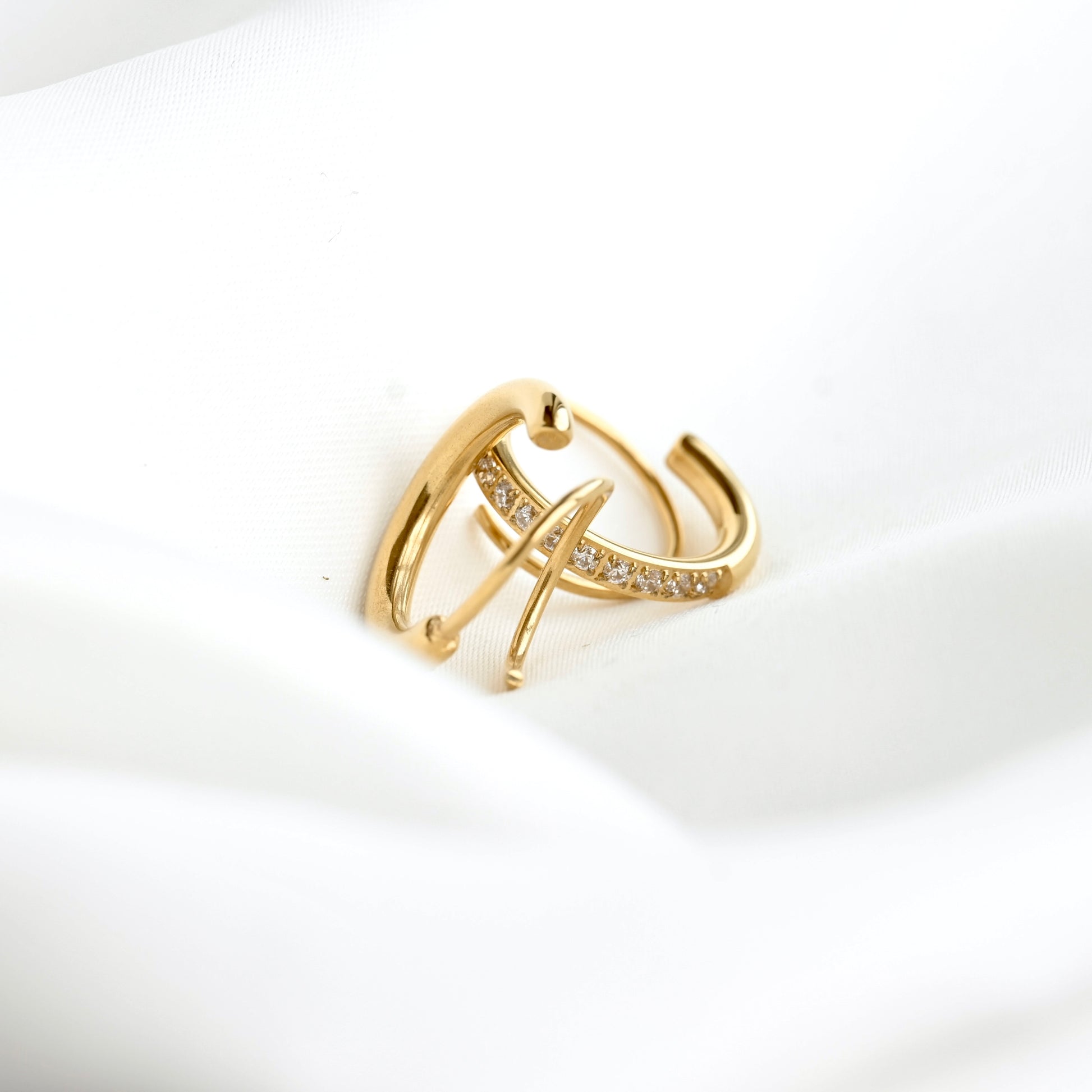 A gold huggie hoop ring with a curved bar studded with small clear crystals, resting on a soft white fabric background.