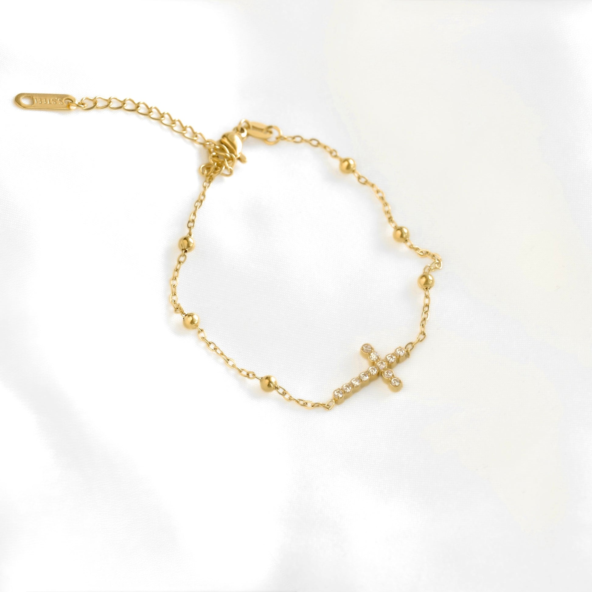 A dainty gold cross bracelet with a beaded chain, horizontally placed cross charm, and a lobster clasp with extender.