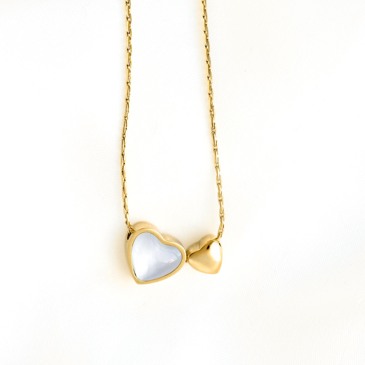 A gold heart pendant necklace featuring one larger heart with a white mother-of-pearl inlay and one smaller gold heart beside it, suspended on a delicate gold chain against a plain white background.