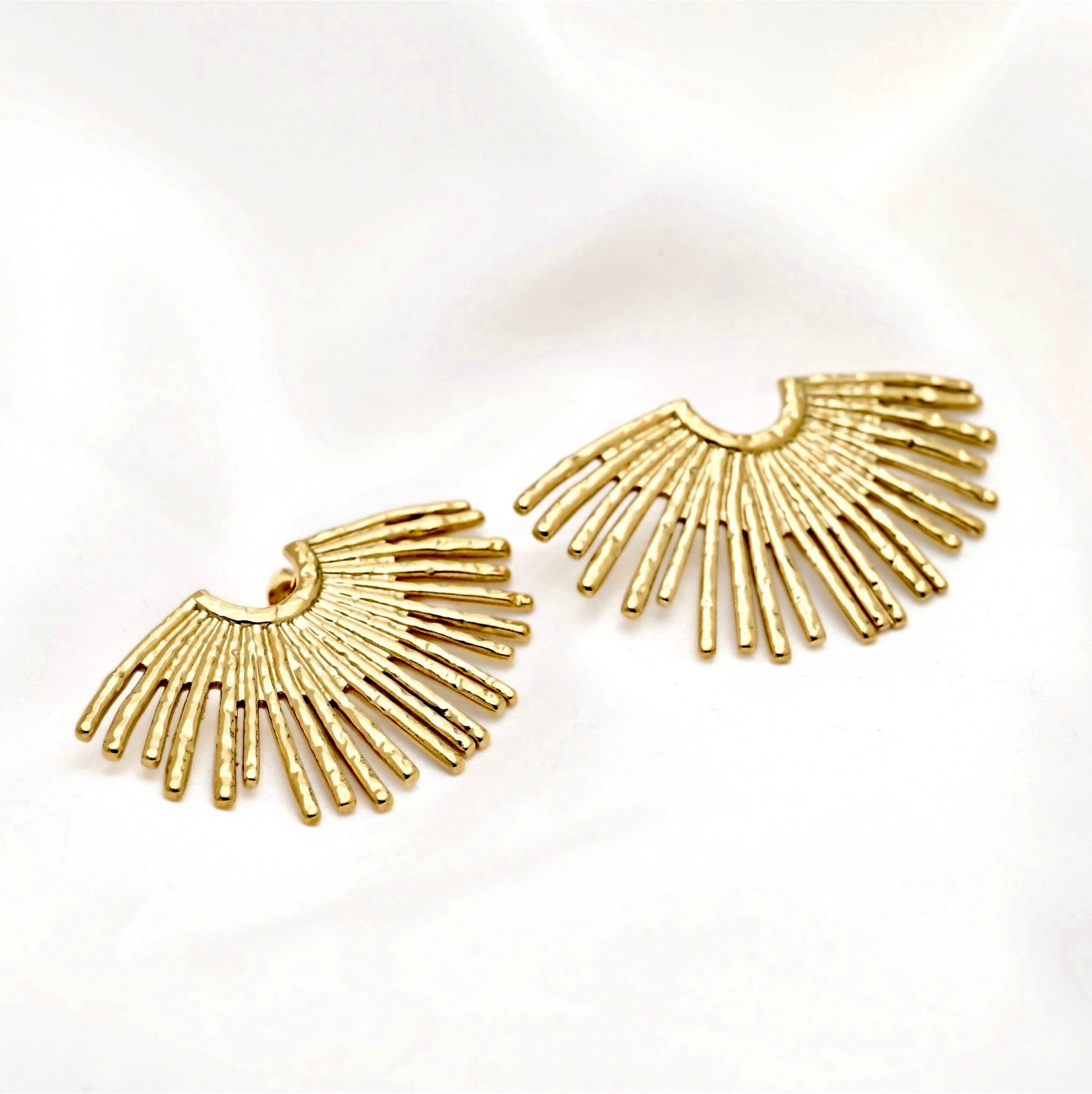 A pair of gold-toned crescent-shaped stud earrings with radiating rod details, displayed on a soft white fabric background.