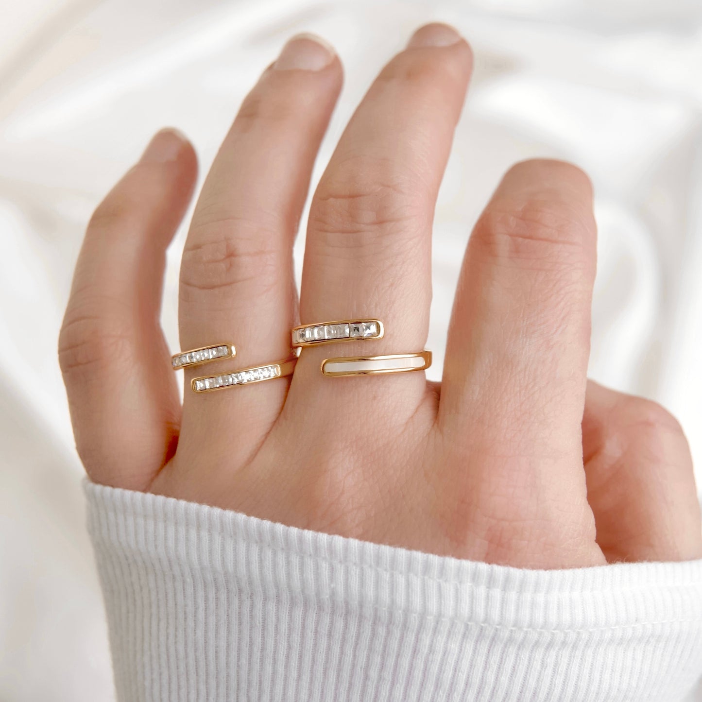 A close-up of a hand wearing two slim gold rings, including one with small clear stones, against a soft white fabric background.