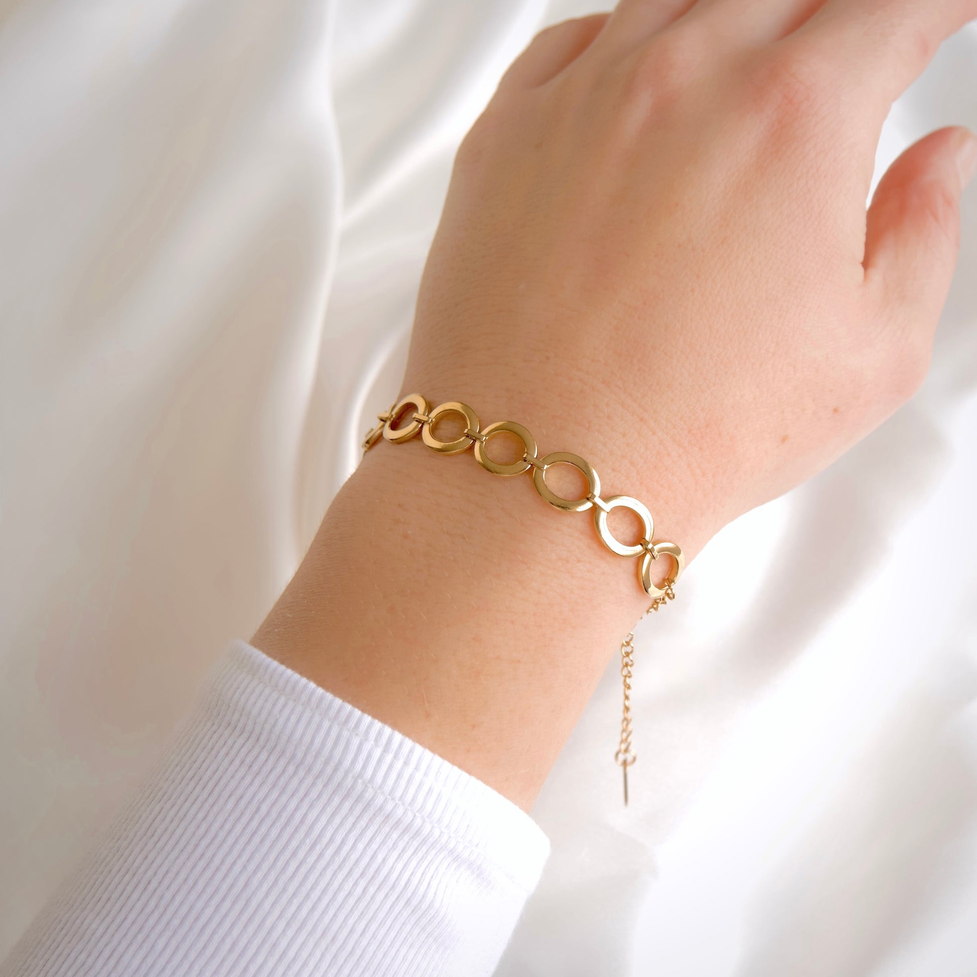 A gold link bracelet with interlocking circular rings worn on a person's wrist, shown against a soft white fabric background.