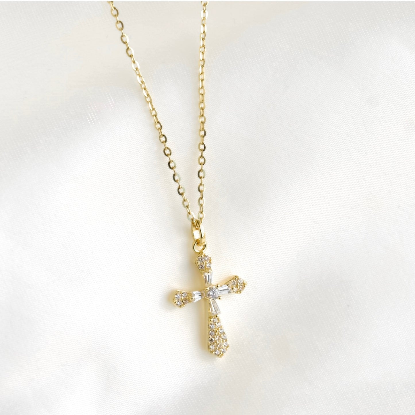 A gold cross pendant necklace adorned with clear crystals, displayed on a soft white fabric background.