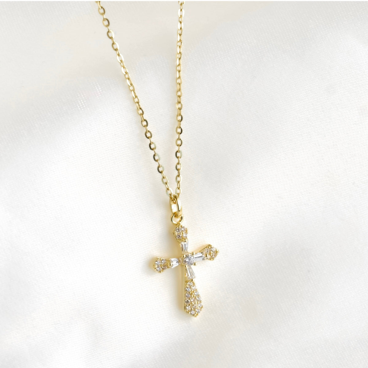 A gold cross pendant necklace adorned with clear crystals, displayed on a soft white fabric background.