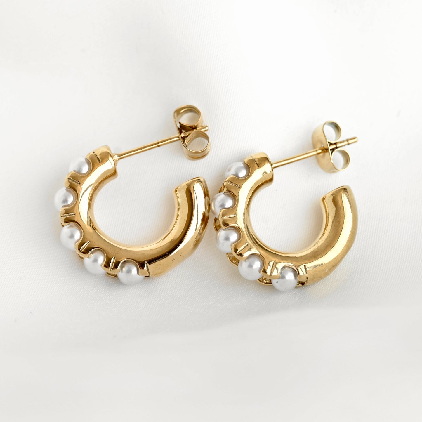 A pair of gold C-shaped hoop earrings decorated with small white pearls along the front edge, displayed on a clean white fabric background.