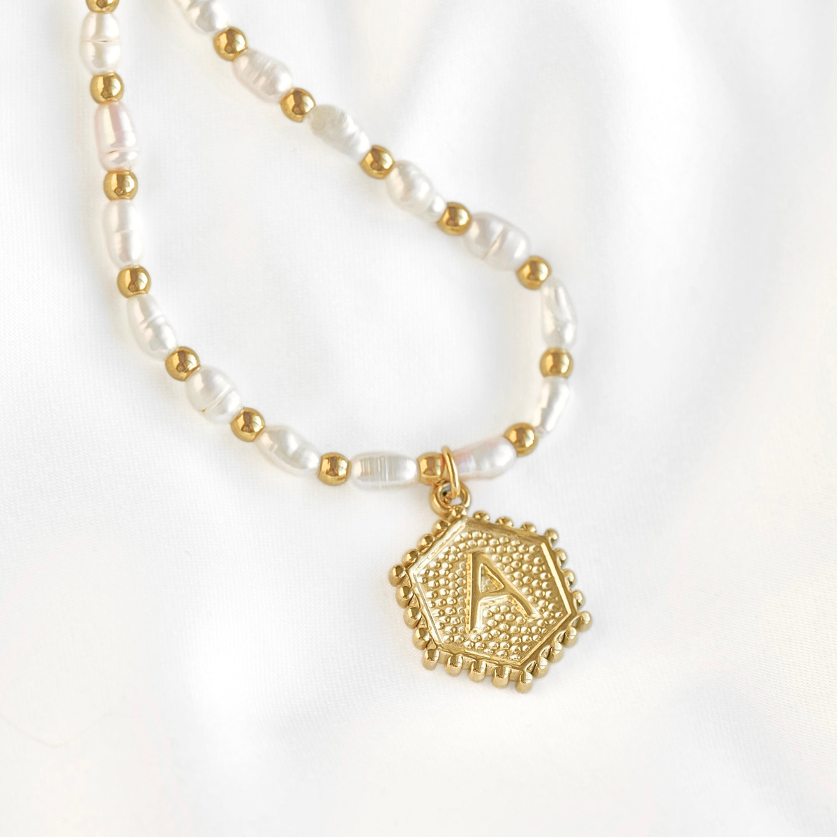 A gold and white beaded necklace featuring irregular white pearl-like beads, small gold spacer beads, and a textured gold hexagonal pendant engraved with the letter 'A', displayed on a soft white fabric background.