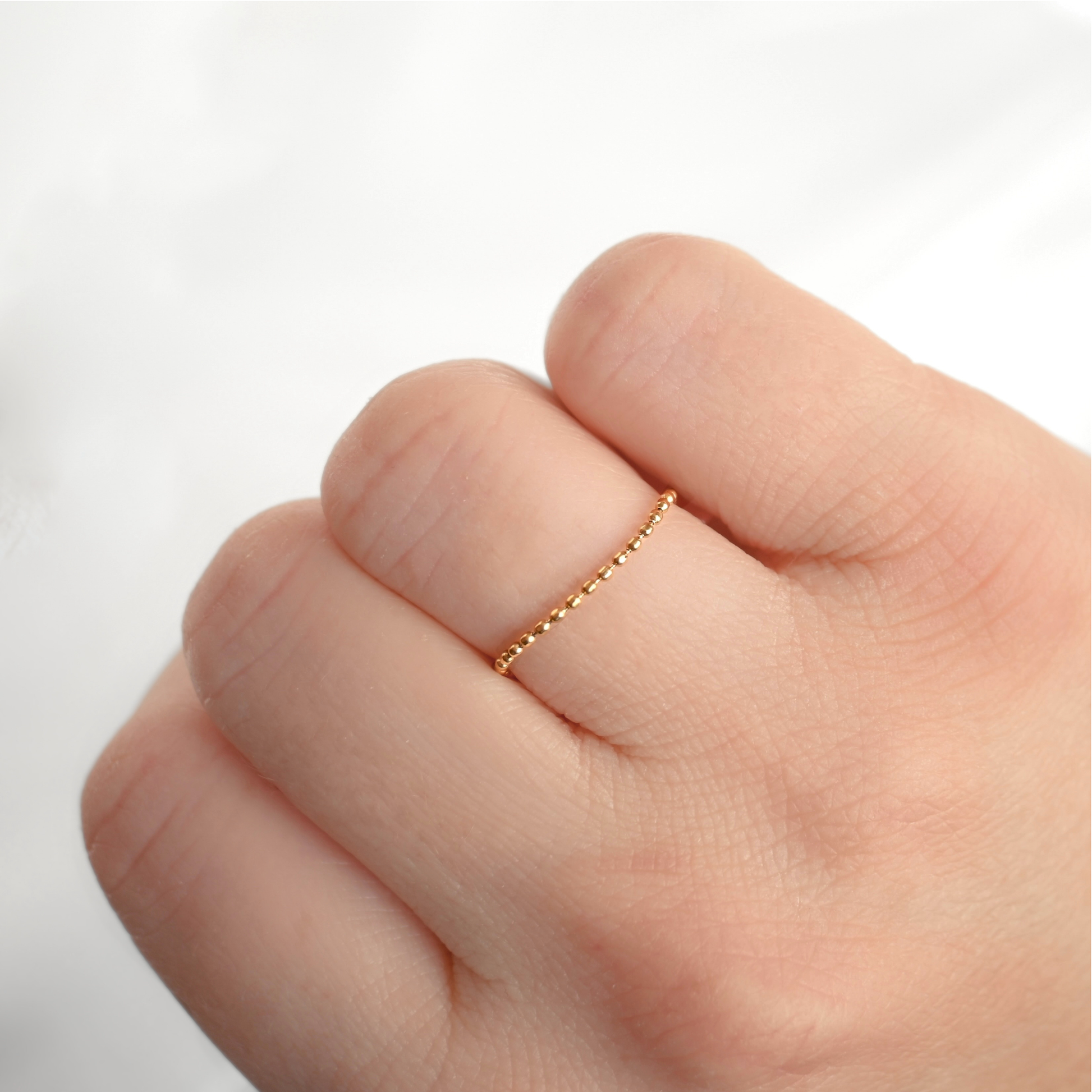 A delicate gold ball chain ring with a small beaded or textured band worn on the ring finger of a hand, shown against a soft, light gray background.