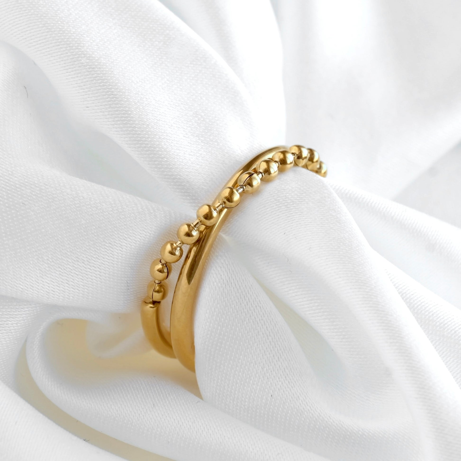 A gold beaded chain criss cross ring with a ball chain crossing oer a smooth gold band on a white draped fabric background.