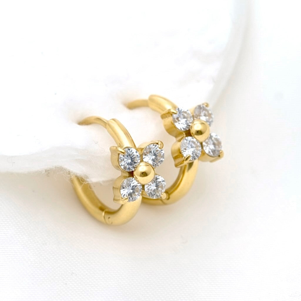 A pair of gold huggie hoop earrings with small flower clusters of clear round-cut stones and central gold beads, displayed on a soft white fabric surface.