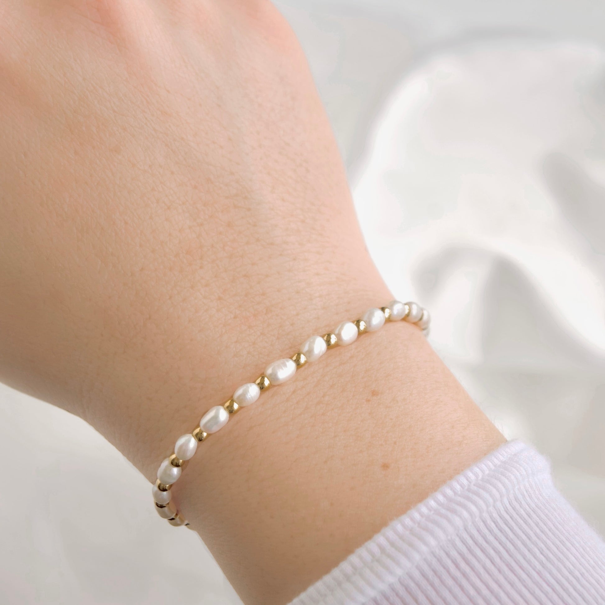 A delicate bracelet with small white pearls and gold spacer beads worn on a person's wrist, shown against a soft white background.