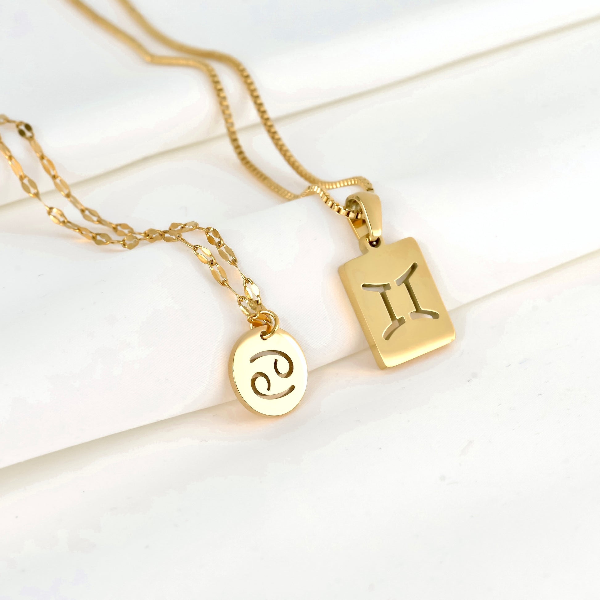 Two gold necklaces with zodiac pendants, one coin and one tablet on a white surface