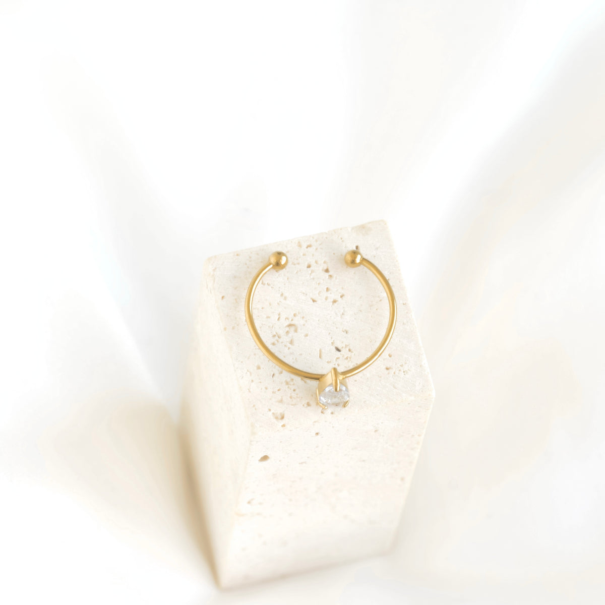A delicate gold hoop earring with a small clear gemstone at the bottom, displayed on a textured cream-colored stone block against a soft white background.