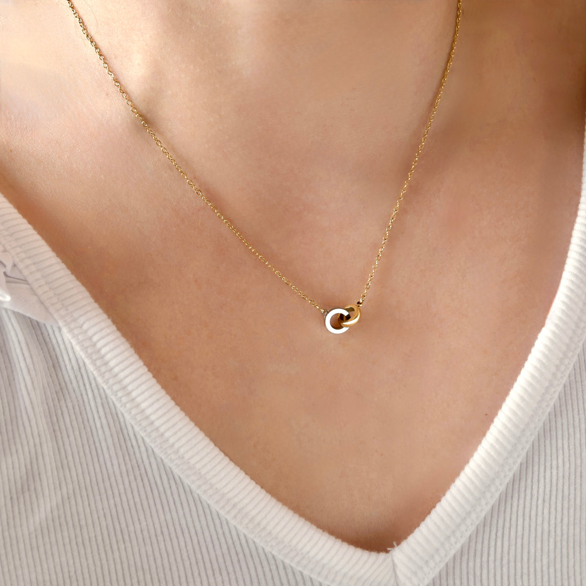 A close-up of a person's neck and chest wearing a delicate gold chain necklace with a small interlocking ring pendant, paired with a white ribbed V-neck top.