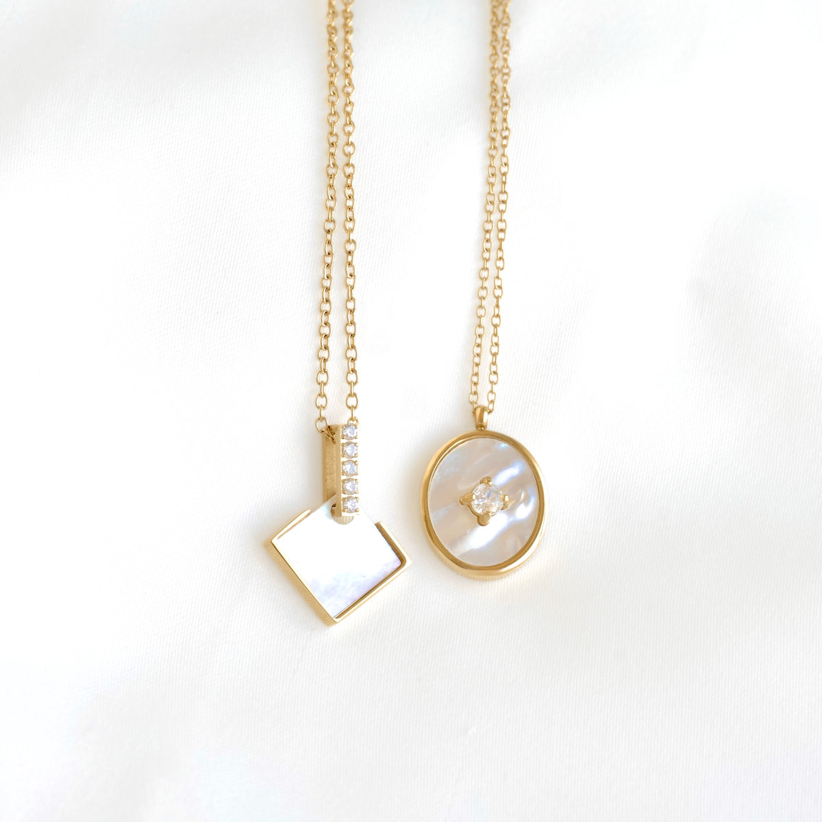 Two gold chain necklaces displayed on a white background. One necklace features a square pendant with a white iridescent inlay and a small vertical row of clear stones.