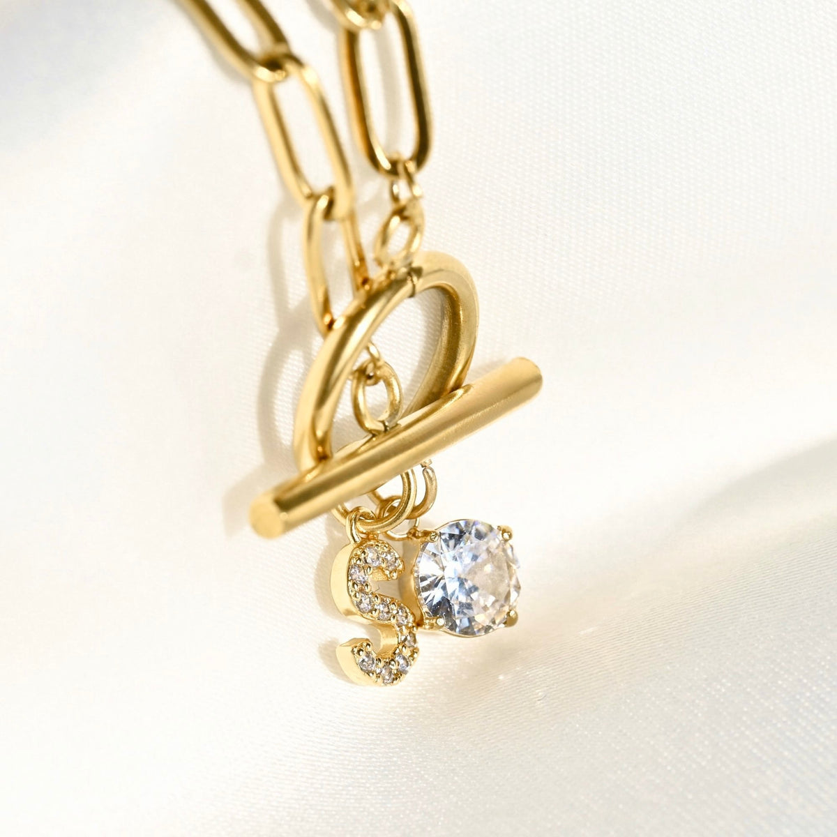 Gold chain necklace with a toggle clasp and a dangling initial 'S' charm embellished with small clear stones, set against a white fabric background.
