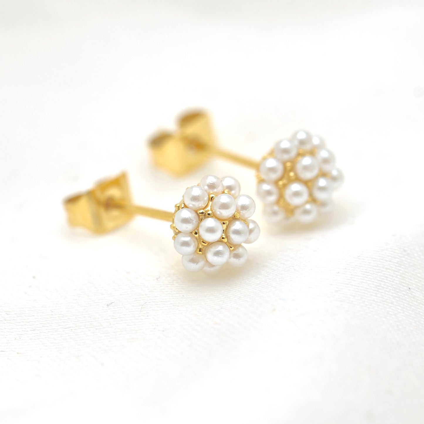 A pair of gold stud earrings with white pearl flower clusters, displayed on a clean white textured fabric background.