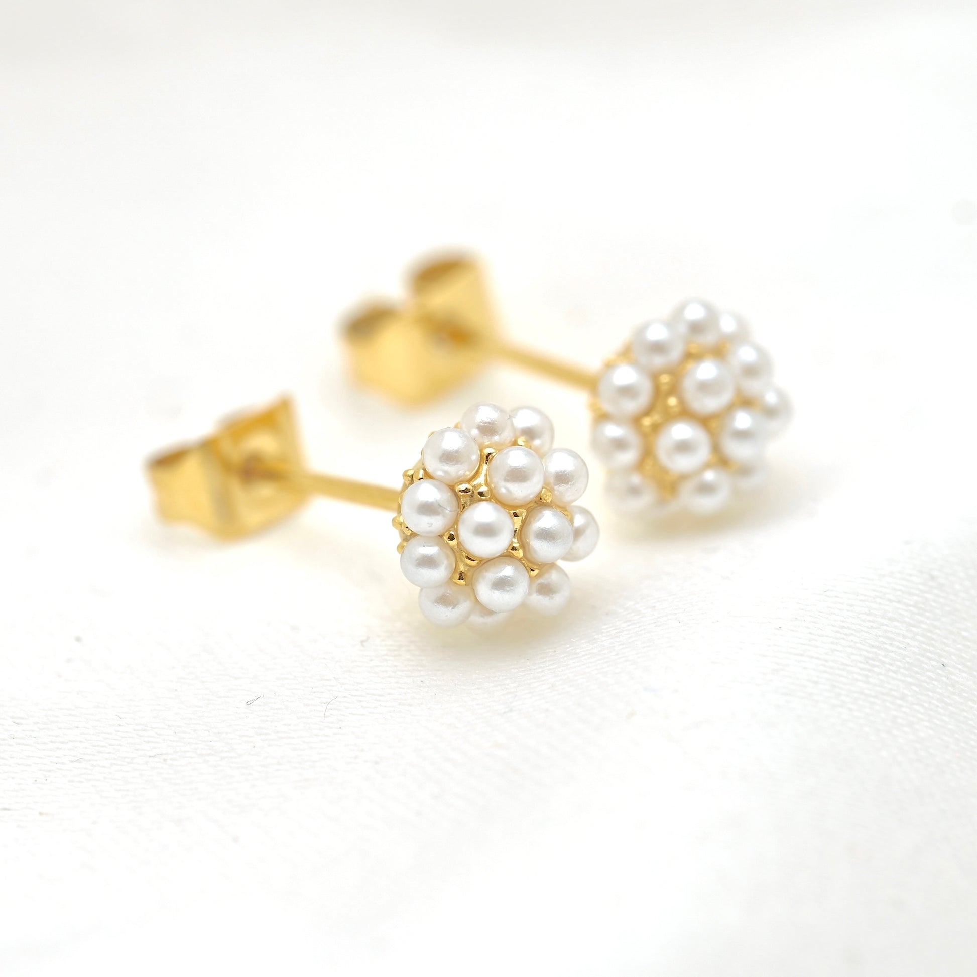 A pair of gold stud earrings with white pearl flower clusters, displayed on a clean white textured fabric background.