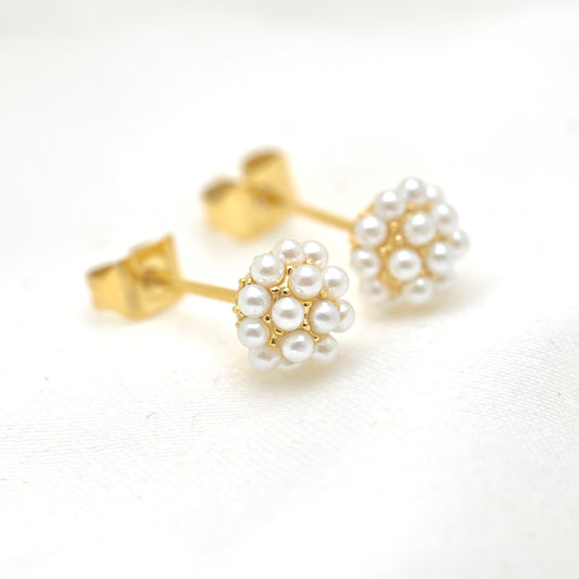 A pair of gold stud earrings with white pearl flower clusters, displayed on a clean white textured fabric background.
