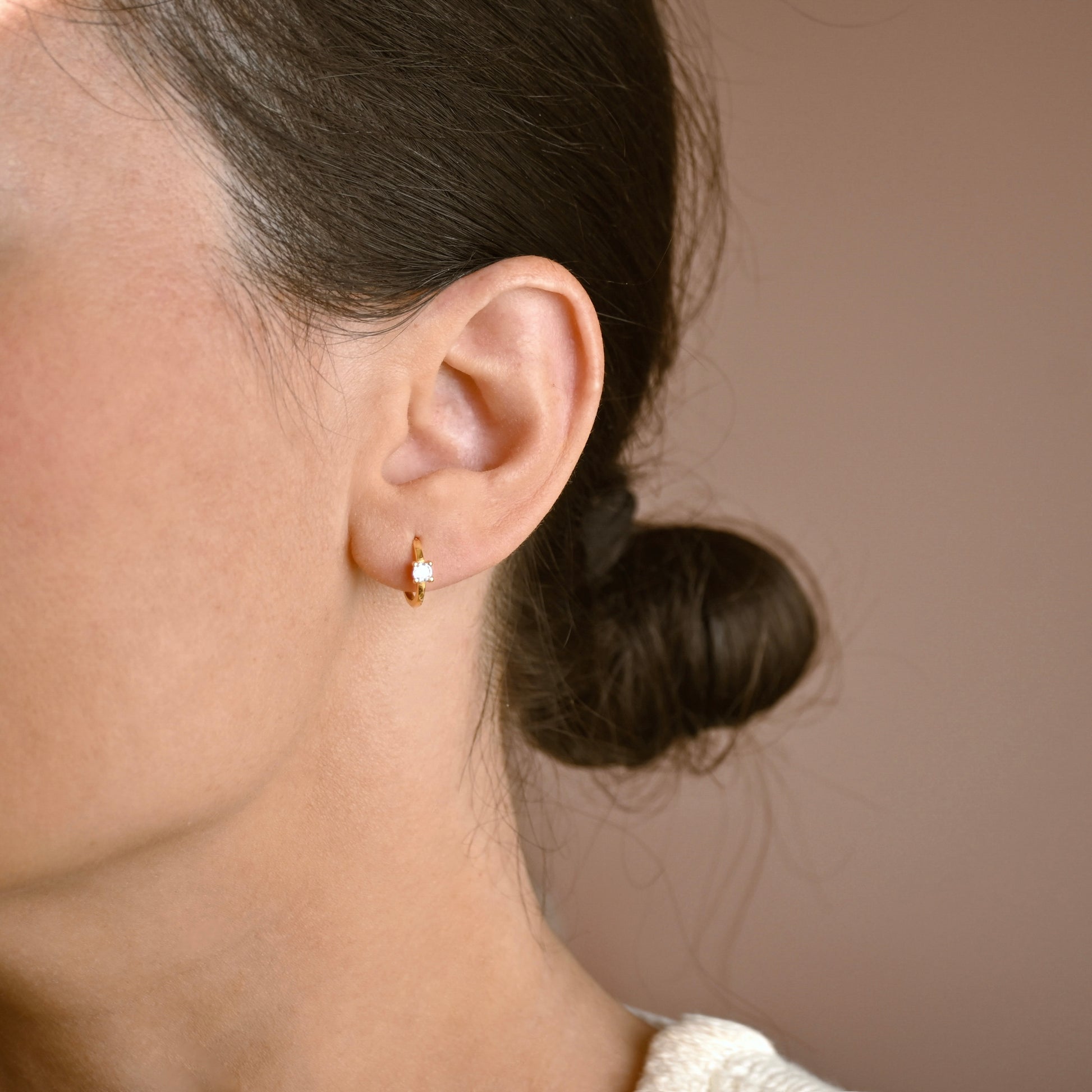 A close-up side profile of a woman wearing a small gold huggie hoop earring with a clear cubic zirconia, set against a solid warm brown background.