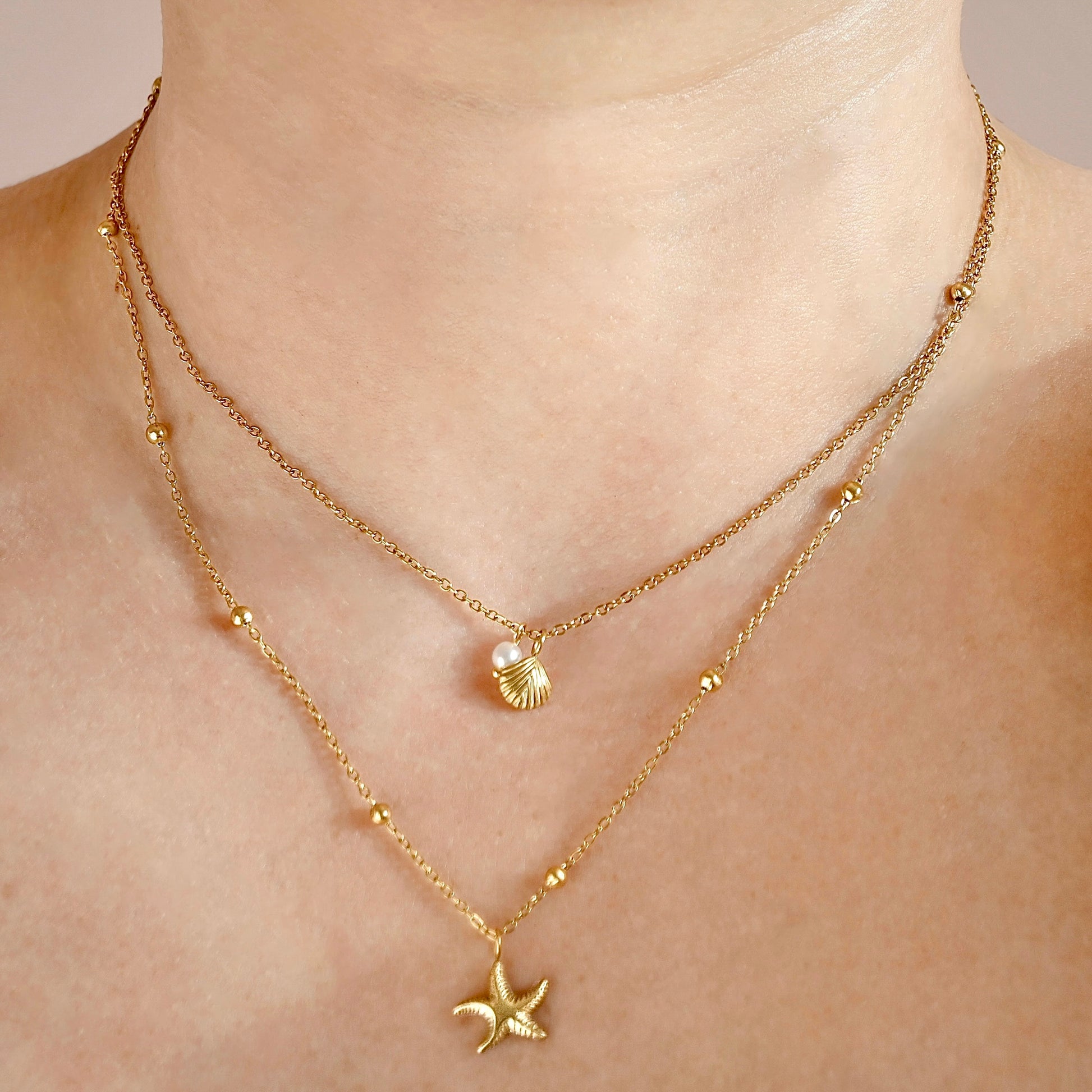 A close-up of a person's neck and upper chest wearing a delicate layered gold necklace set. The top layer features a small seashell pendant with a pearl accent, and the longer bottom layer features a gold starfish pendant, both accented with small gold beads.