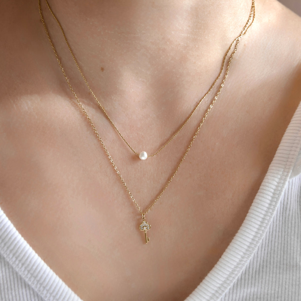 A close-up of a person's neck and chest wearing a delicate double-layered gold necklace. The top layer is a fine chain with a small white pearl, and the bottom layer has a small key-shaped pendant.