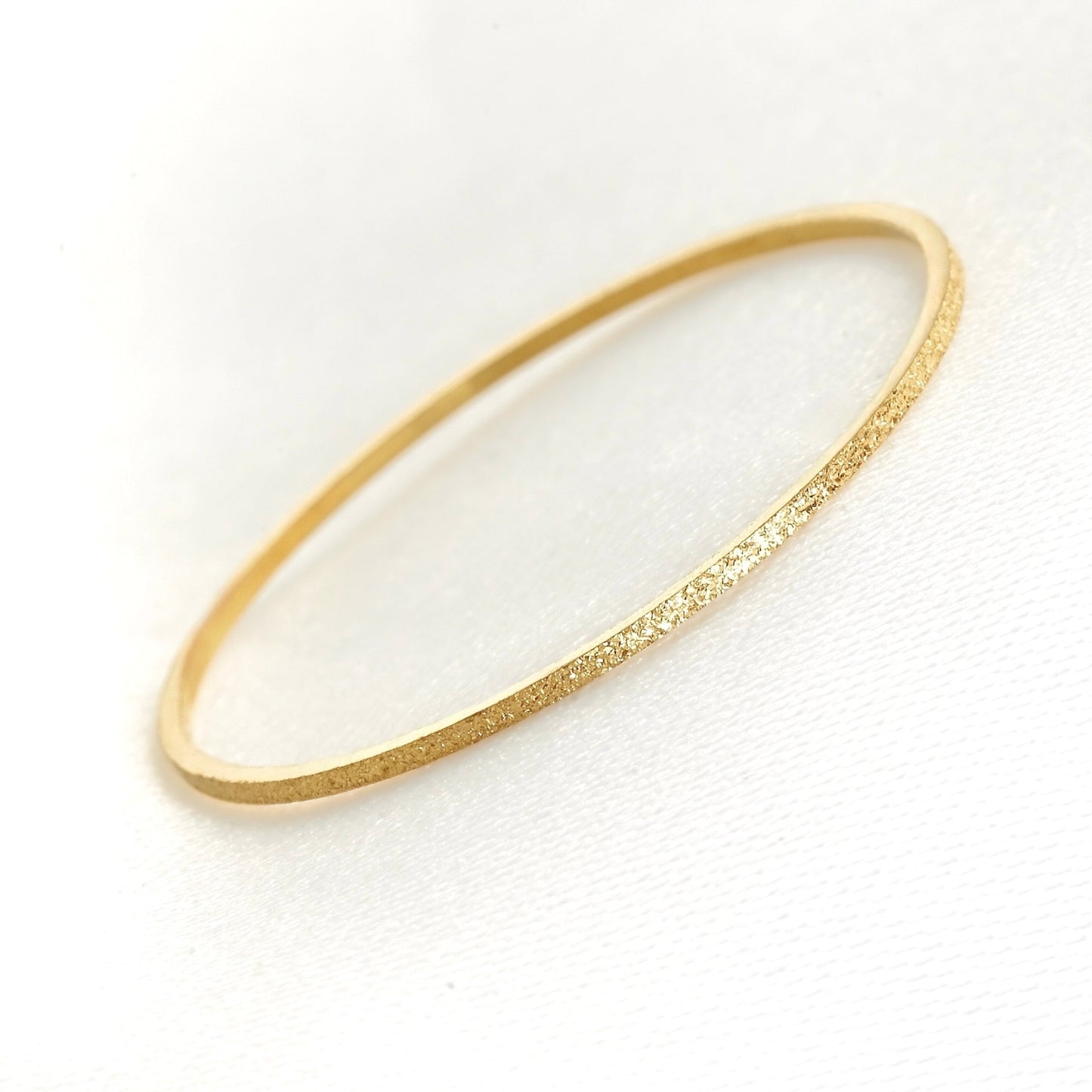 A delicate, thin gold bandwith a textured, glittery finish, displayed on a clean white textured background.