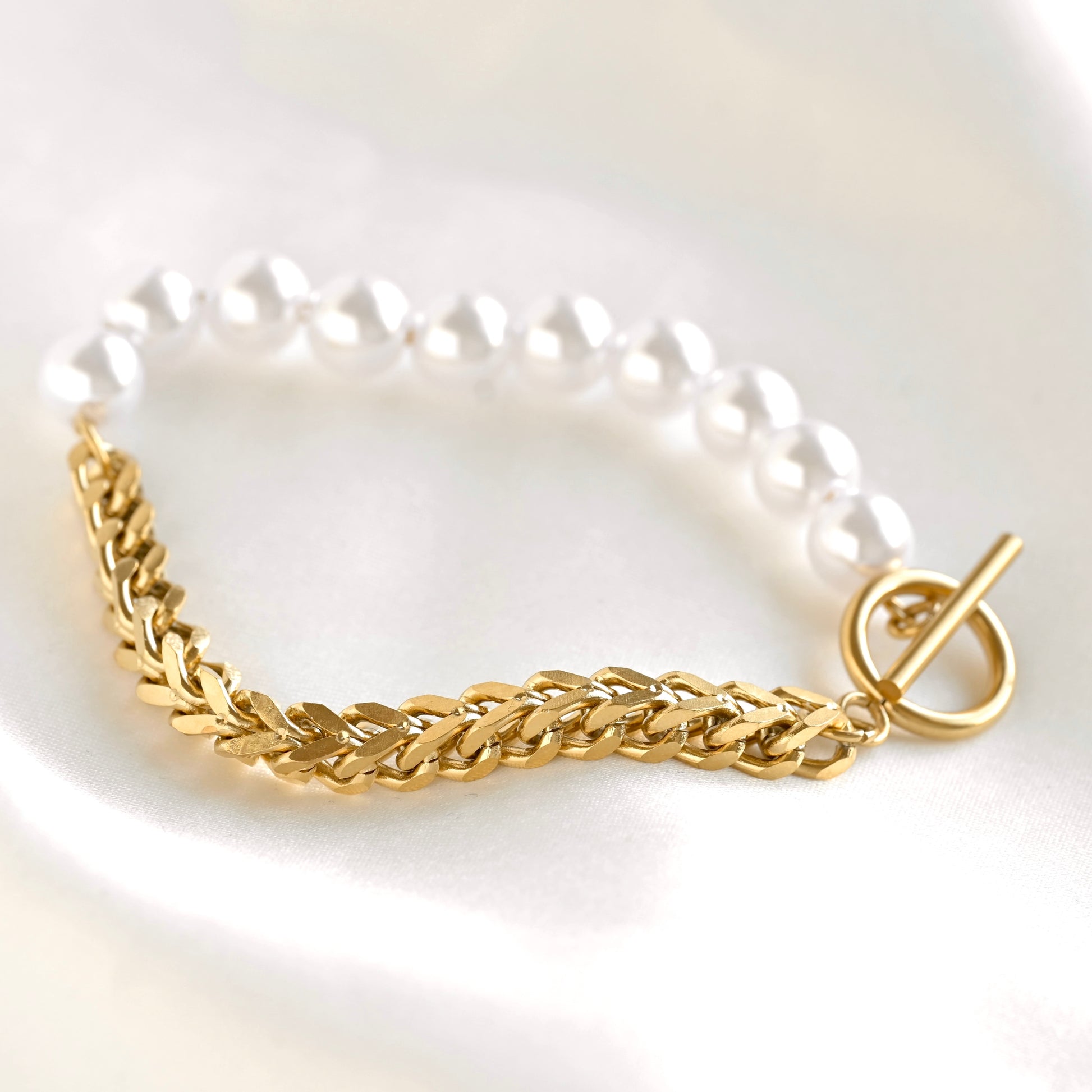 A gold chain bracelet with a braided gold section and a white pearl strand, shown on a soft white background.