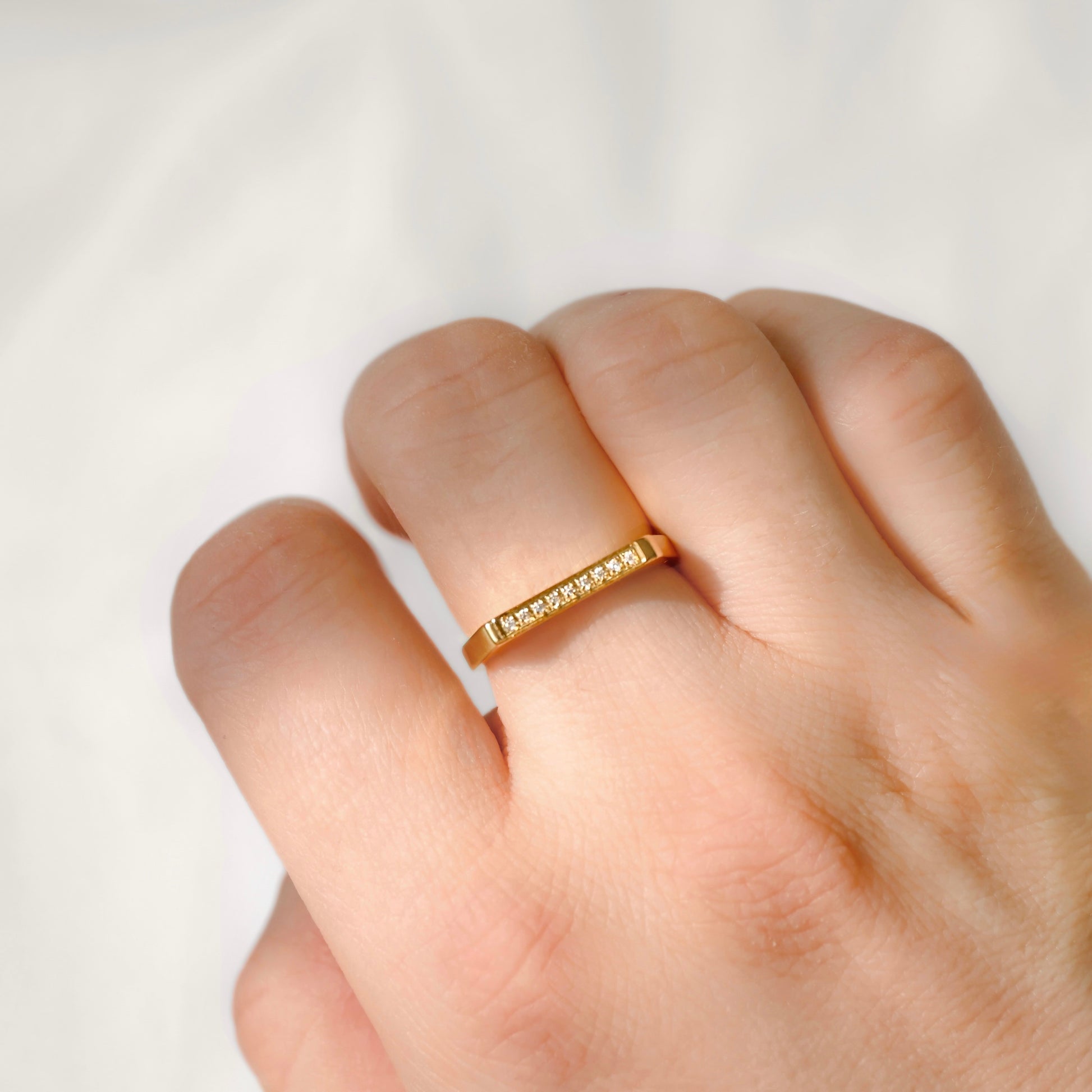 A close-up of a hand wearing a thin yellow gold band ring with a row of small clear stones, set against a soft white fabric background.