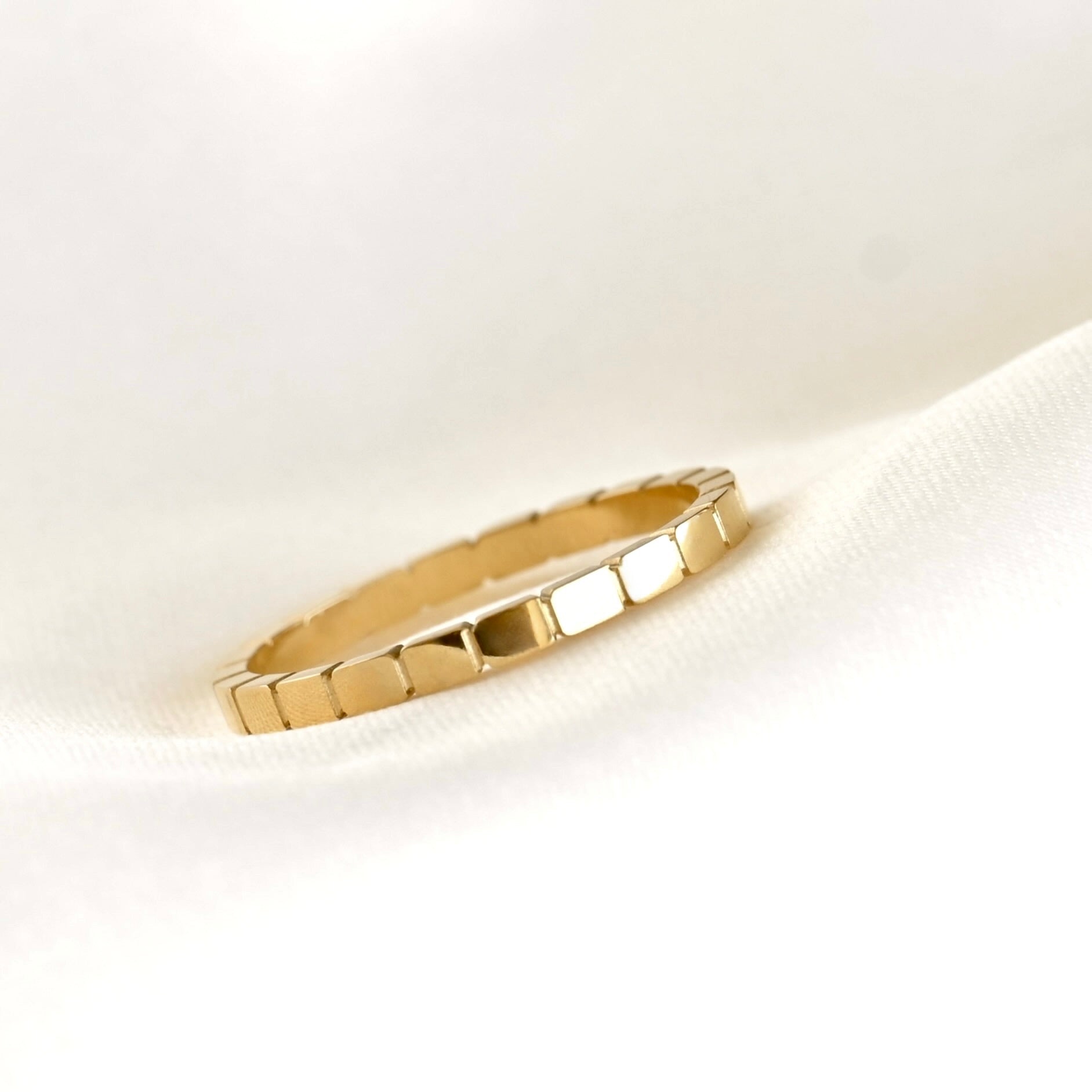 A gold ring with a textured, block-like segmented design, resting on a soft white fabric background.
