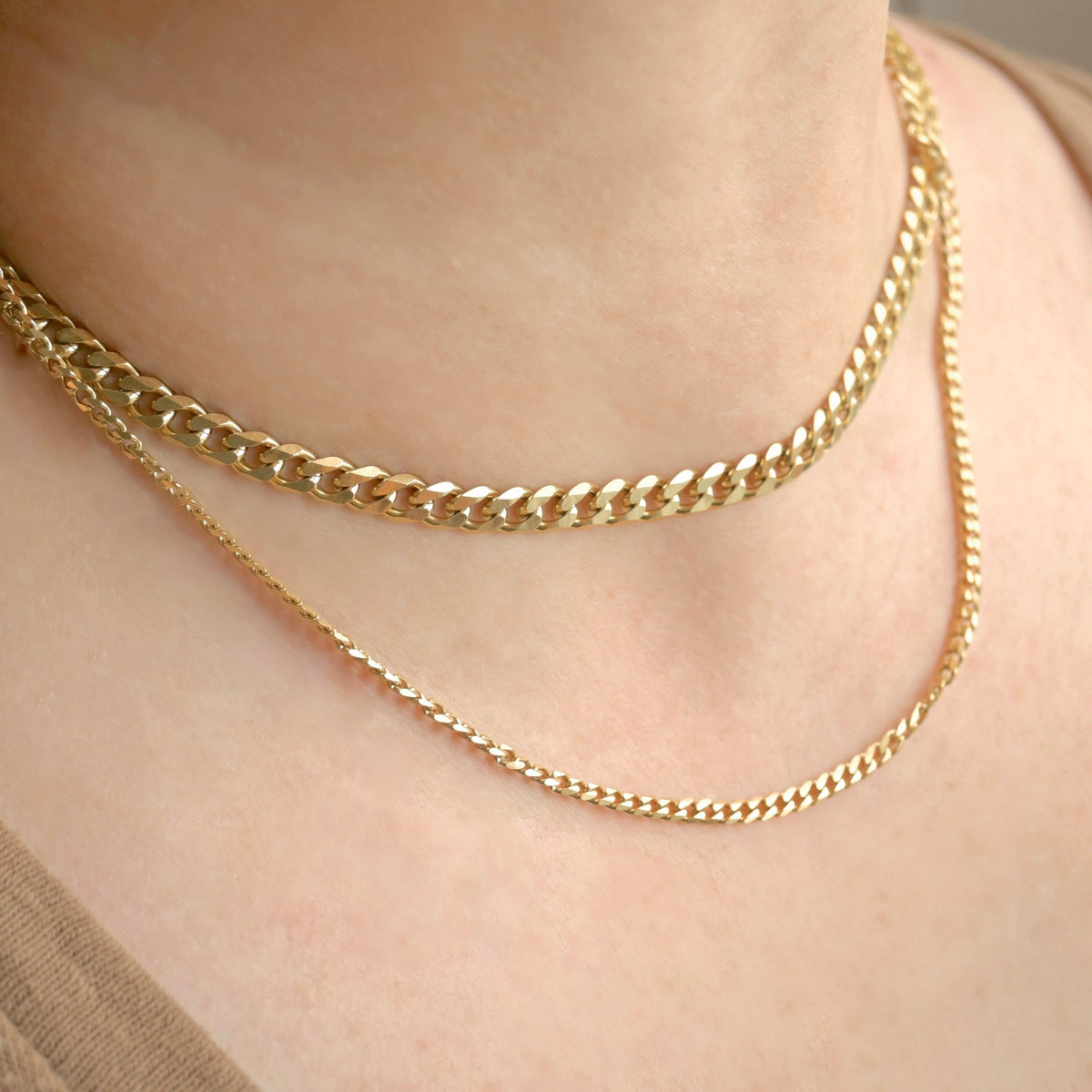 Two layered gold-tone necklaces worn on a person's neck: a thicker curb chain on top and a finer chain below, shown against light skin.