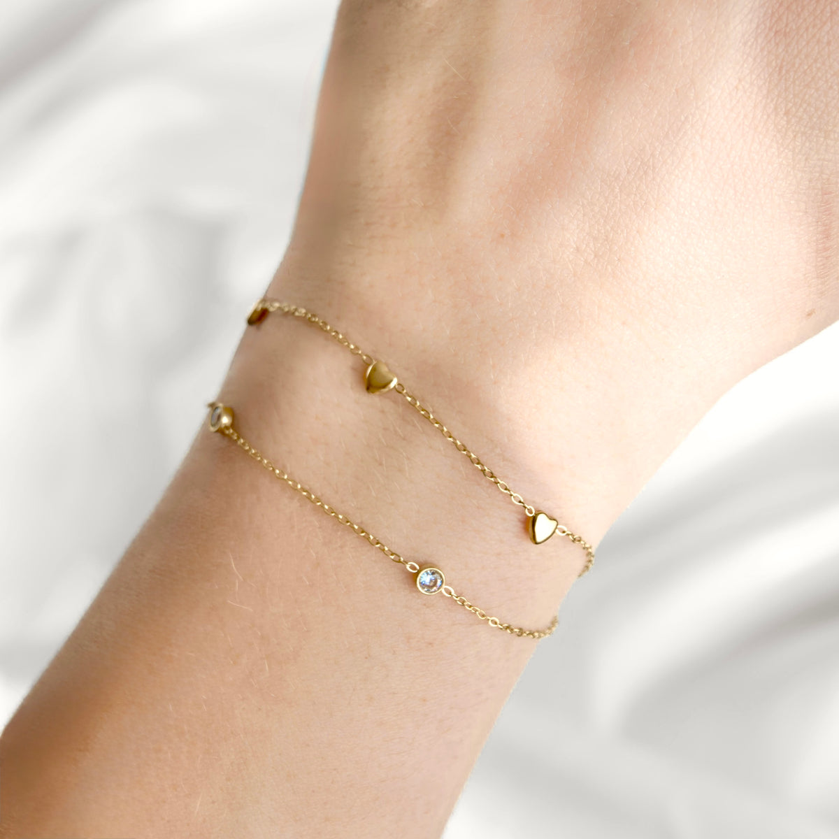 Two delicate gold chain bracelets with small heart charms and a tiny clear stone, shown worn on a person's wrist against a soft white background.