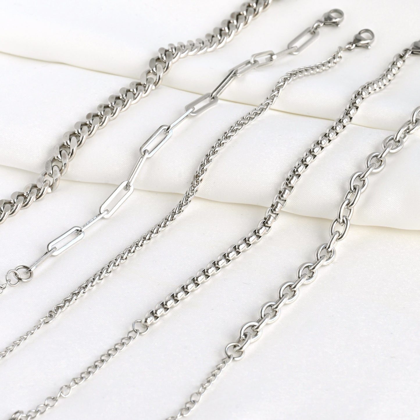 Silver chain bracelets on a light satin fabric. From left to right: Curb, paperclip, wheat, rounded box and rolo