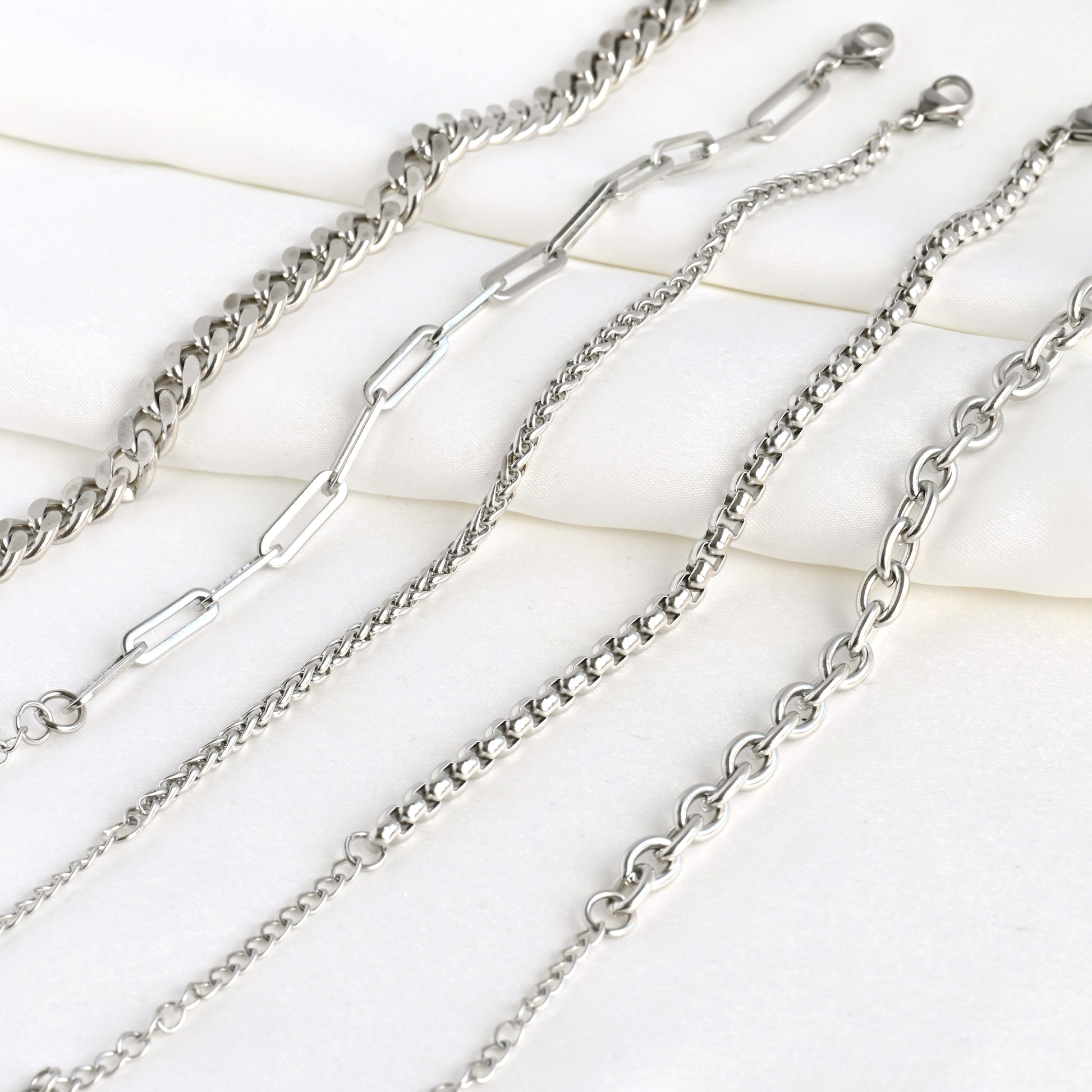 Silver chain bracelets on a light satin fabric. From left to right: Curb, paperclip, wheat, rounded box and rolo