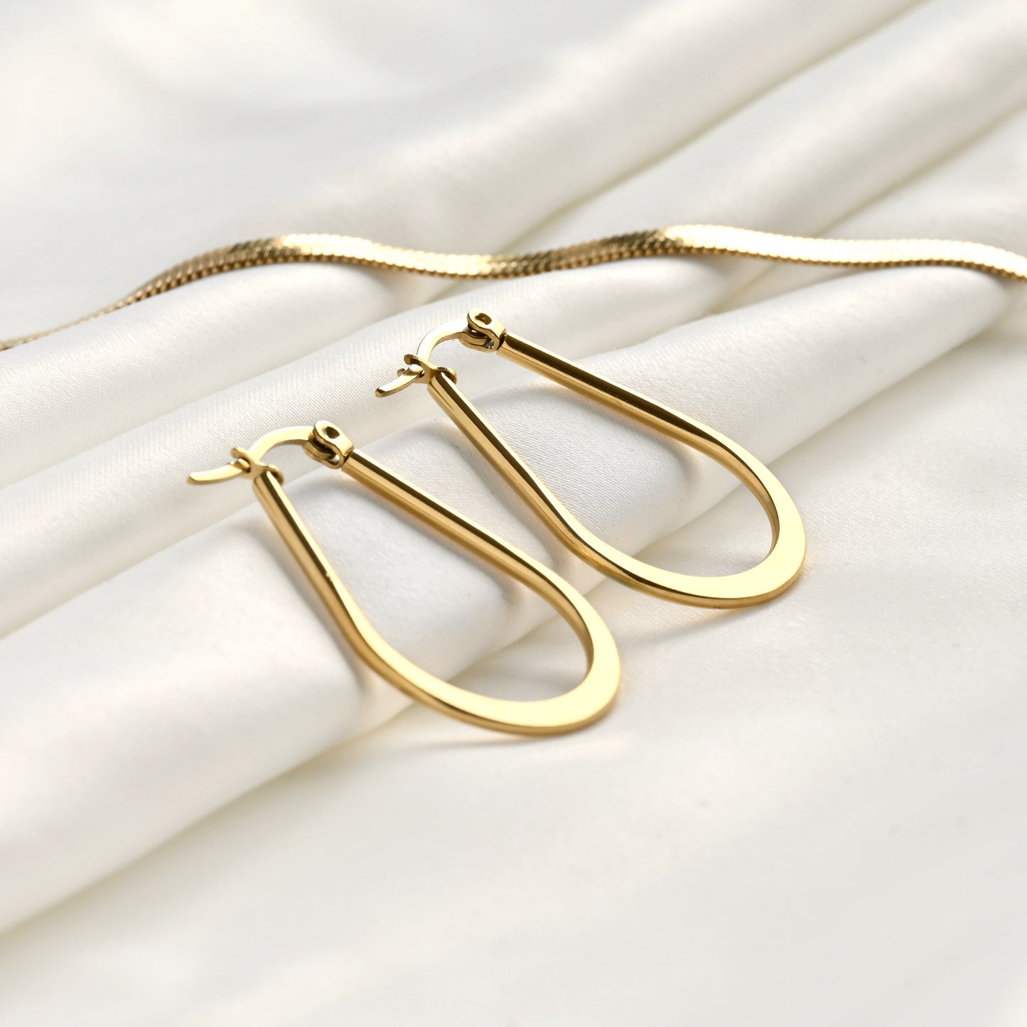 Gold teardrop hoop earrings on a white fabric background with a gold herrringbone chain at the back