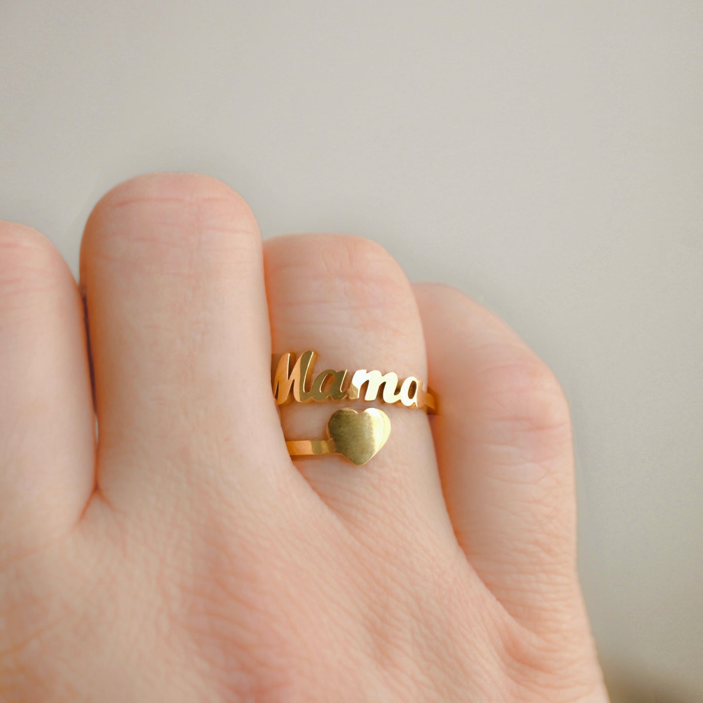 A gold ring worn on a finger, featuring the word 'Mama' in cursive script and a small heart, shown on a hand against a soft neutral background.
