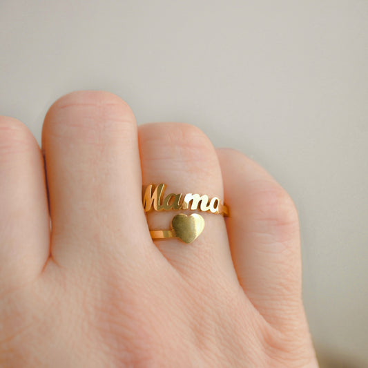 A gold ring worn on a finger, featuring the word 'Mama' in cursive script and a small heart, shown on a hand against a soft neutral background.