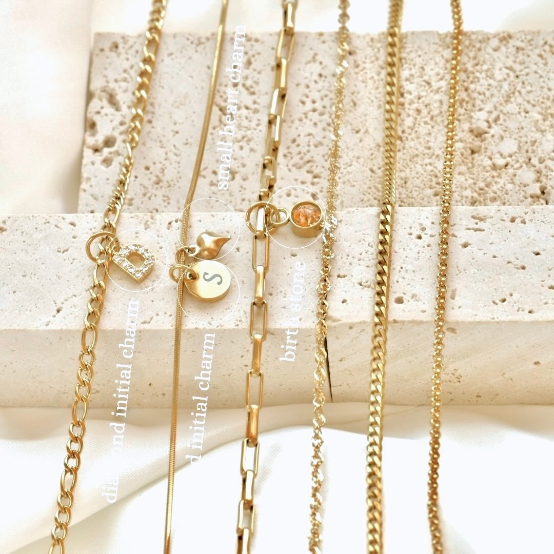 Various gold chain necklaces with initial, heart and birthstone charms on a textured surface