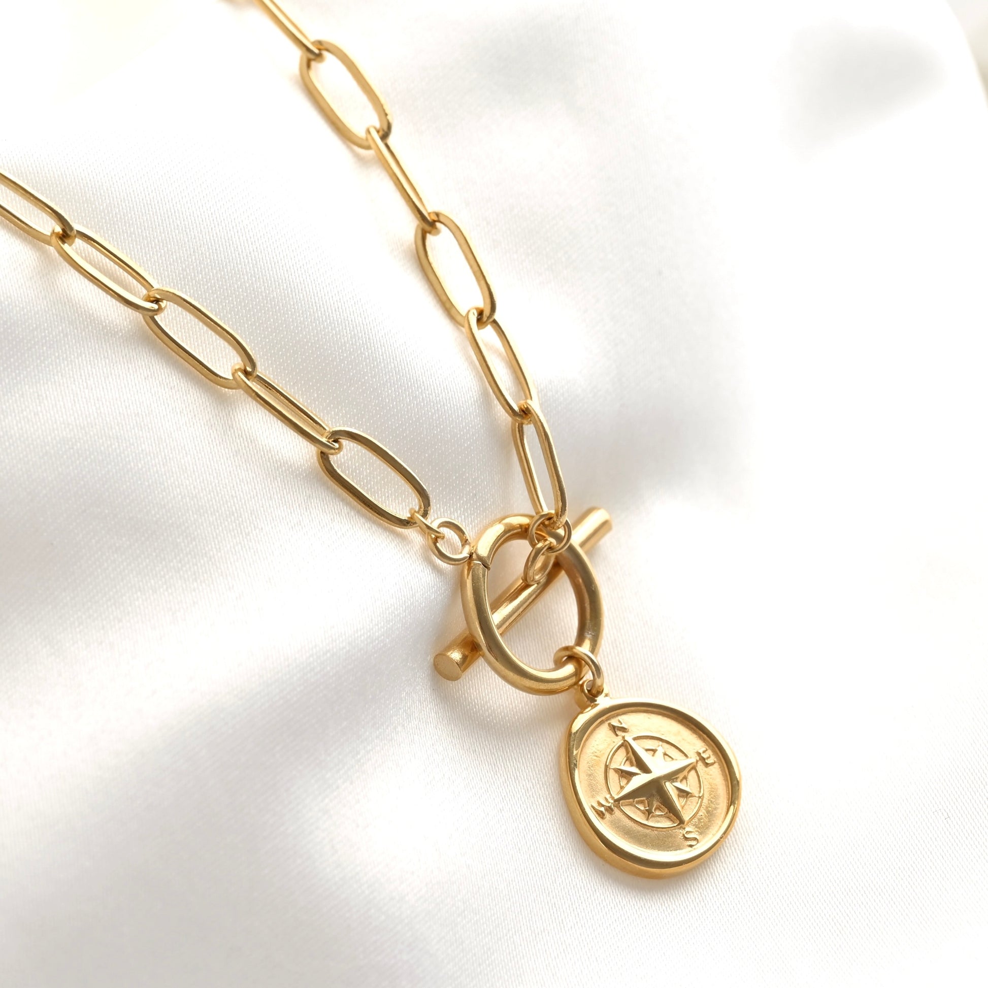 A gold-toned paperclip chain necklace with a toggle clasp and a round compass pendant, displayed on a white satin fabric background.