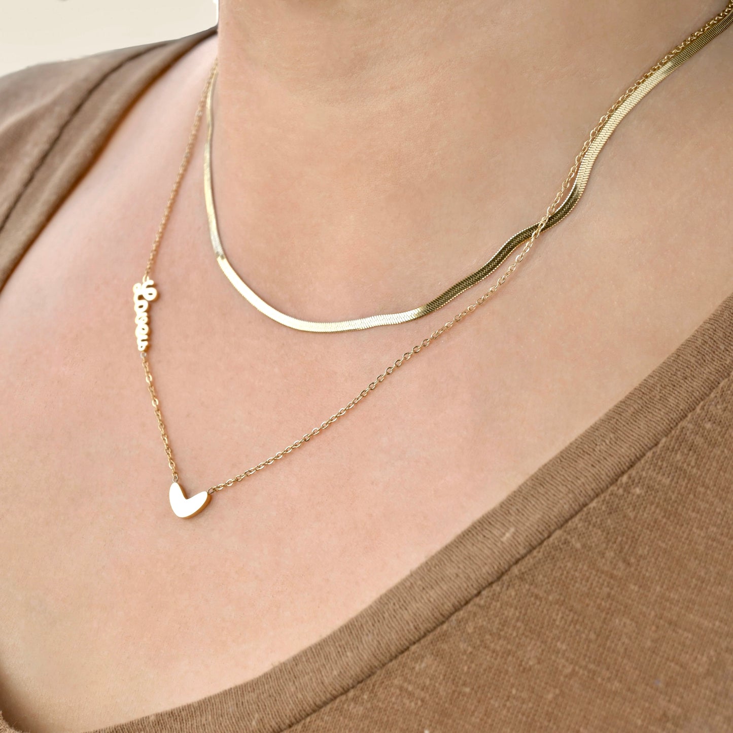 Gold layered herringbone and cable chain necklace with a love script and heart charm on a person wearing a brown top