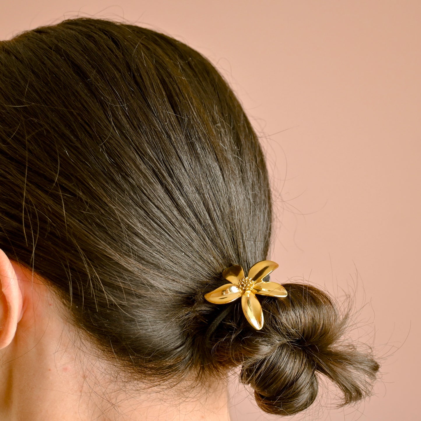 A close-up of a person's hair styled in a low bun, secured with a shiny gold flower-shaped hair tie against a solid warm beige background.