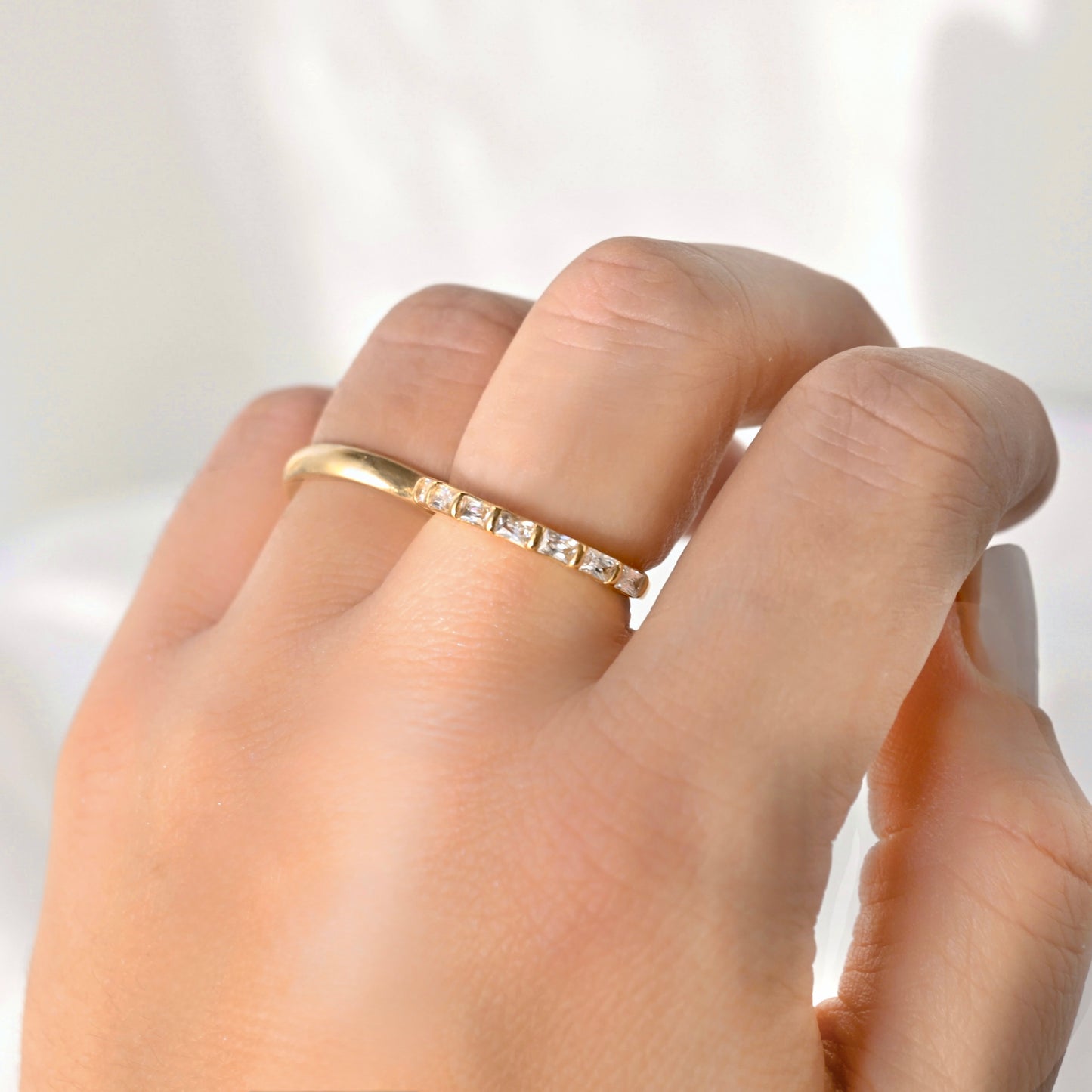 A close-up of a hand wearing a delicate yellow gold band ring with a row of small clear square-cut stones, set against a soft, bright white background.