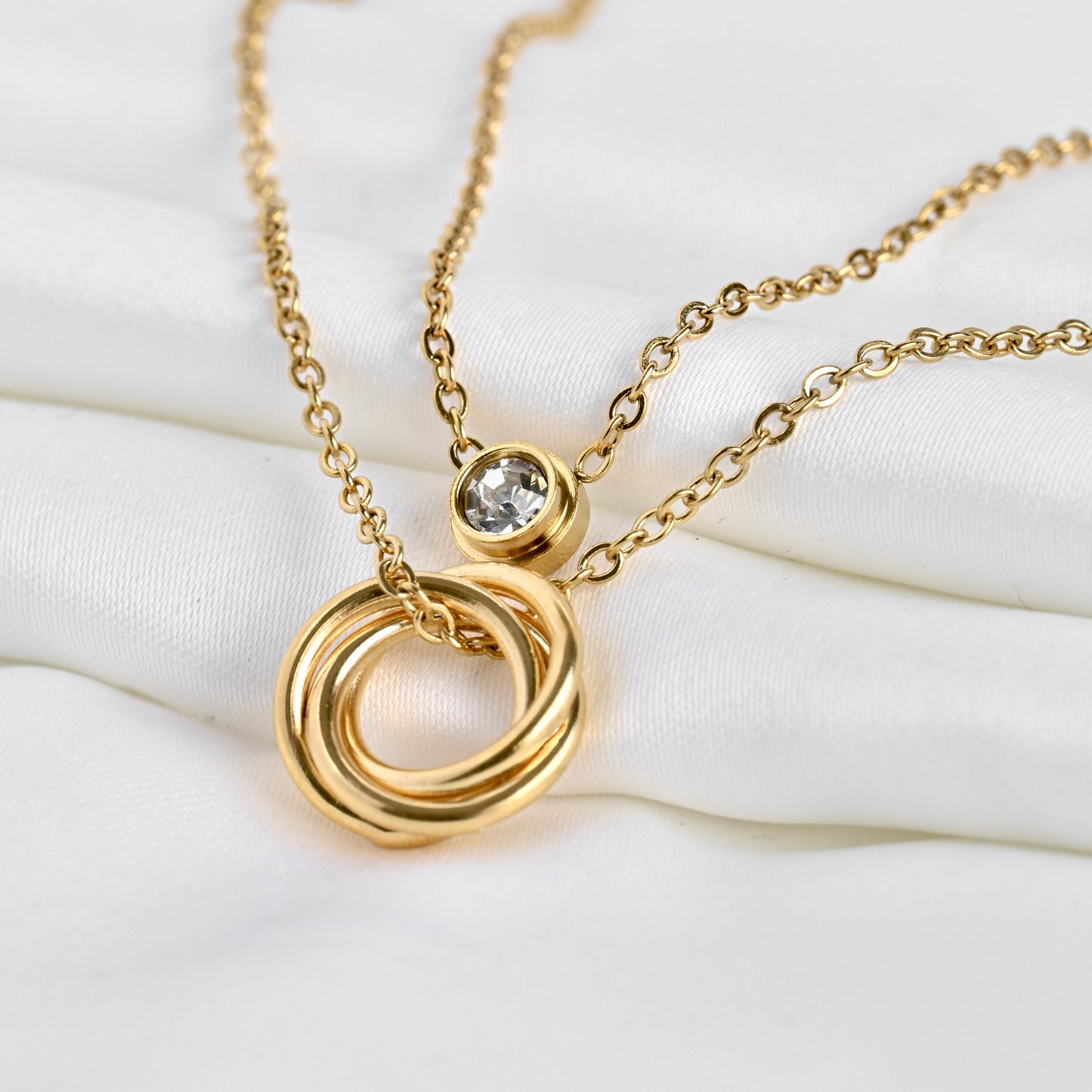 Close up view of double layered gold cable chains, one with an eternity circles pendant, one with bezel crystal on a white surface