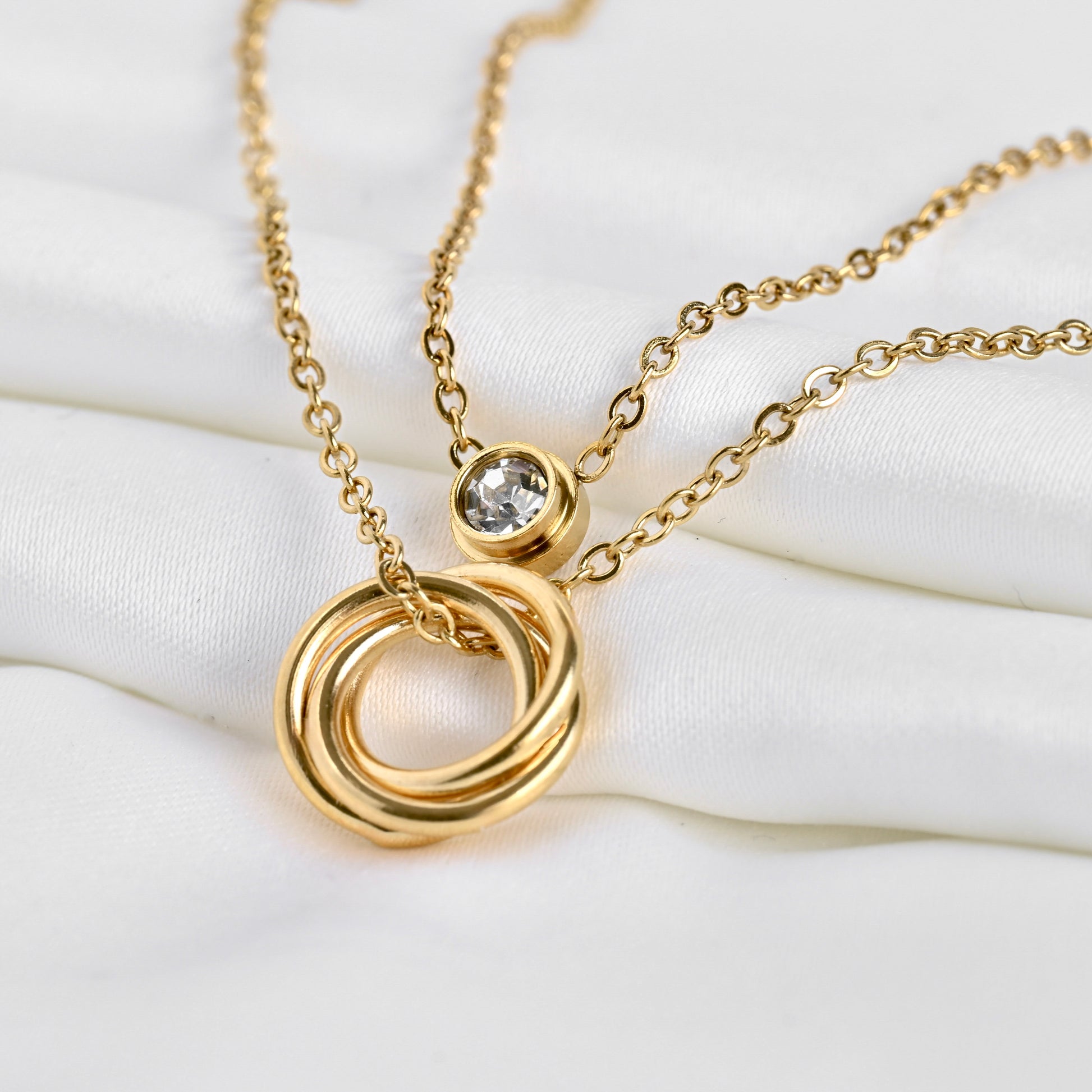 Close up view of double layered gold cable chains, one with an eternity circles pendant, one with bezel crystal on a white surface
