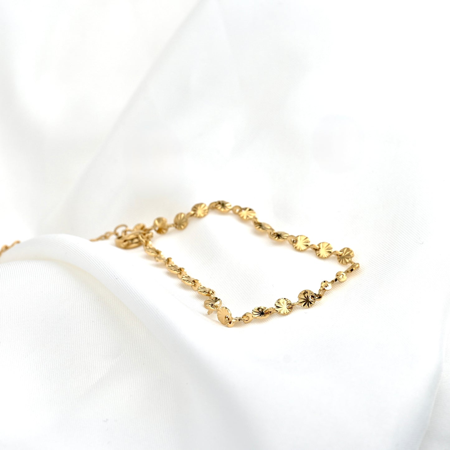 A delicate gold chain bracelet with round sunburst links, displayed on a soft white fabric background.