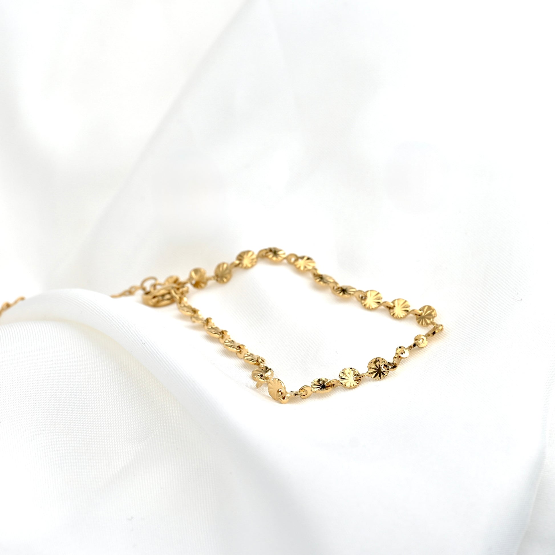 A delicate gold chain bracelet with round sunburst links, displayed on a soft white fabric background.