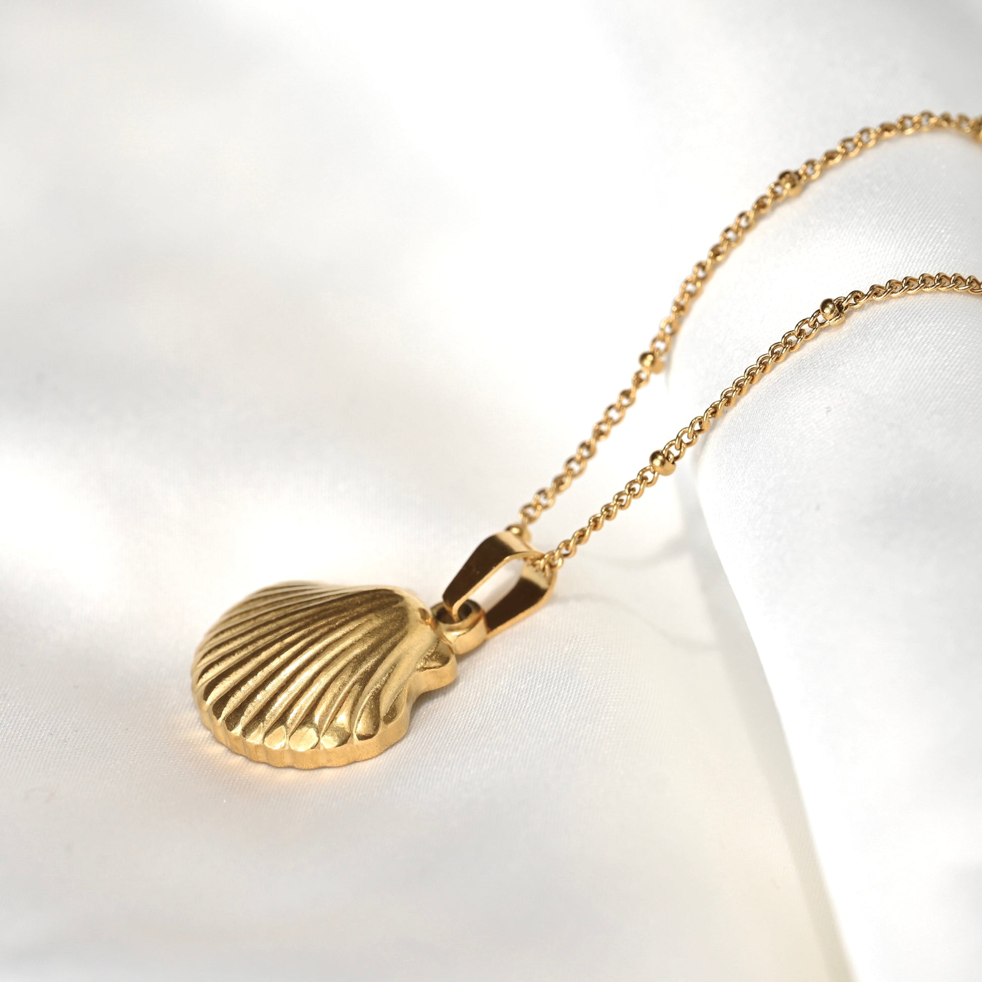 A gold shell pendant necklace with a textured, ribbed scallop shell design, hanging from a delicate gold chain on a soft white fabric background.