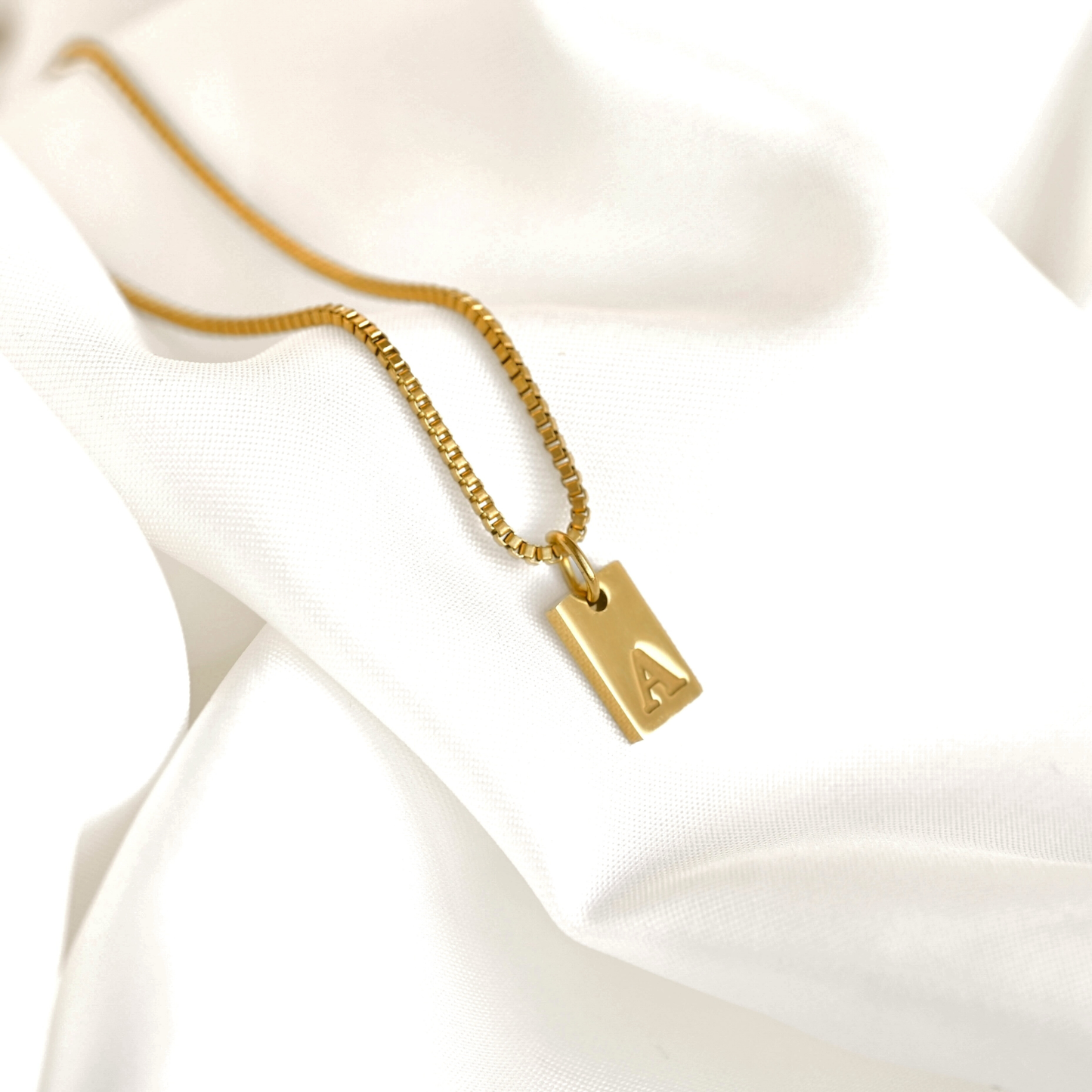 Gold box chain necklace with a rectangular initial pendant on a white satin background